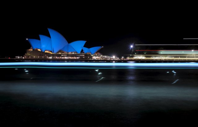 Ferries create long exposure streaks of light as they sail past the Sydney Opera House illuminated in a shade of blue